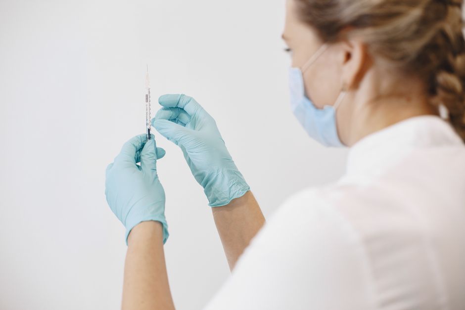 A healthcare worker in gloves and a mask prepares a syringe against a white background.